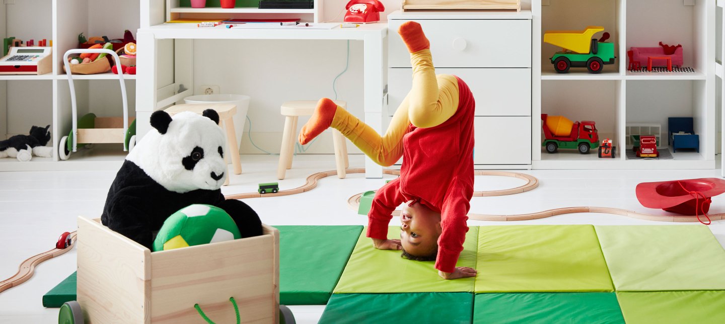 A small child is doing a handstand in a playroom filled with colorful toys and plush animals.