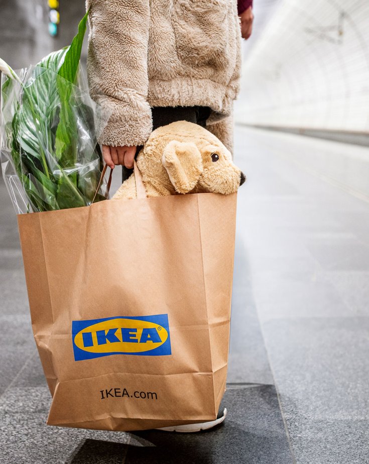A person on a train platform, holding an IKEA paperbag with a plant and a plush toy.