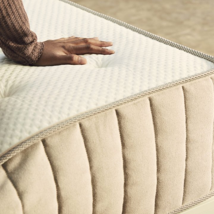 A womans hand pressing against a beige mattress while she sits on it.