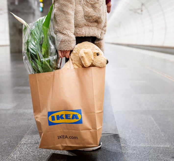 A person on a train platform, holding an IKEA paperbag with a plant and a plush toy.