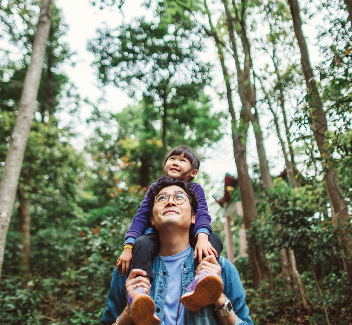 A father carrying his child on his shoulders, standing in a forrest.